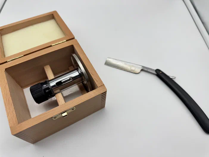 Un microtome manuel rangé dans sa boîte en bois avec son rasoir à côté sur fond blanc.